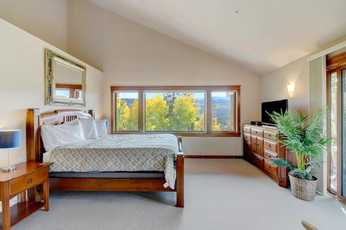 A serene bedroom with an elegant wooden bed facing a large window that frames nature views, complemented by a decorative mirror, modern television, and touches of greenery, creating a soothing space for morning coffee or relaxing at sunrise.