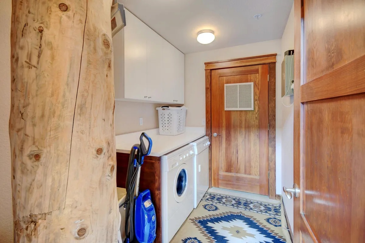 The room features a washer and dryer, a wooden door, a woven rug, and a substantial wooden post. This functional, transitional space is ideal for handling laundry and storing or organizing outdoor gear after a day of adventure.