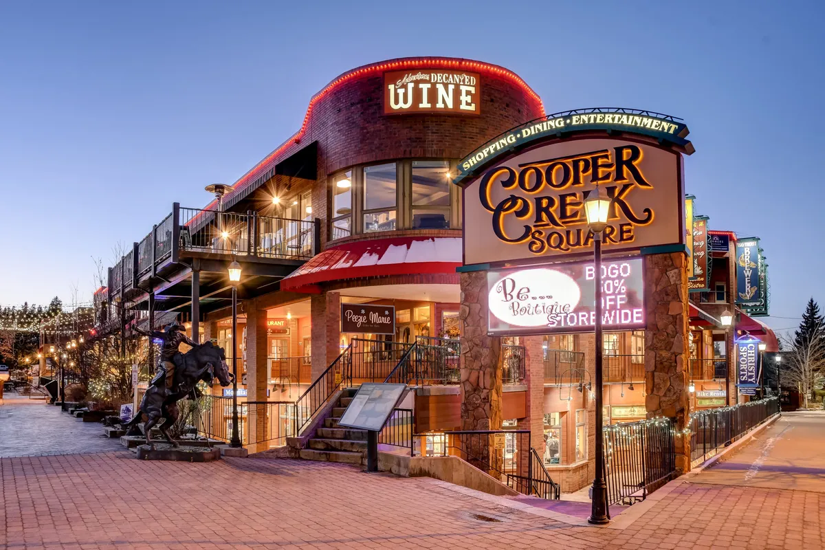 An outdoor view of Cooper Creek Square, featuring a lively shopping and dining hub with a prominent sign, a wine shop, an upper-level staircase, and a horse statue. People are seen engaging with boutique shops and restaurants in an inviting atmosphere.
