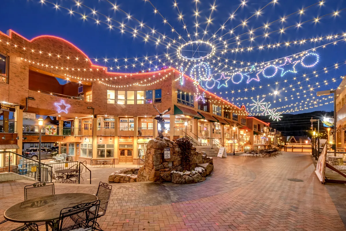 A lively outdoor community square adorned with string lights, decorative storefronts, a central fountain, and ample seating. The space is bustling with opportunities for al fresco dining, shopping, and festive events, suggesting a vibrant local area for families to explore together.