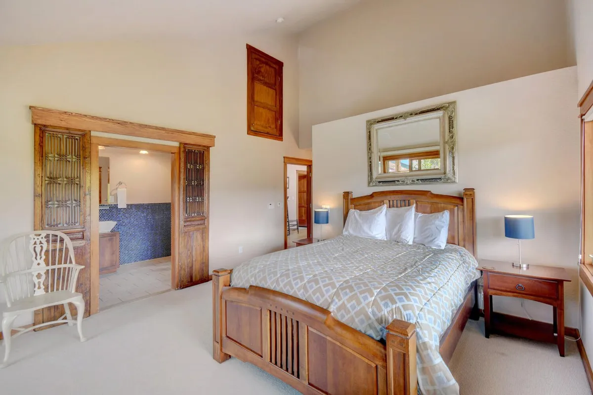 A serene bedroom featuring a wooden bed frame topped with plush bedding, blue accent lamps on matching nightstands, a decorative mirror, and a woven chair in the corner. Gentle natural light fills the space, highlighting the room’s tranquil, cozy mood.