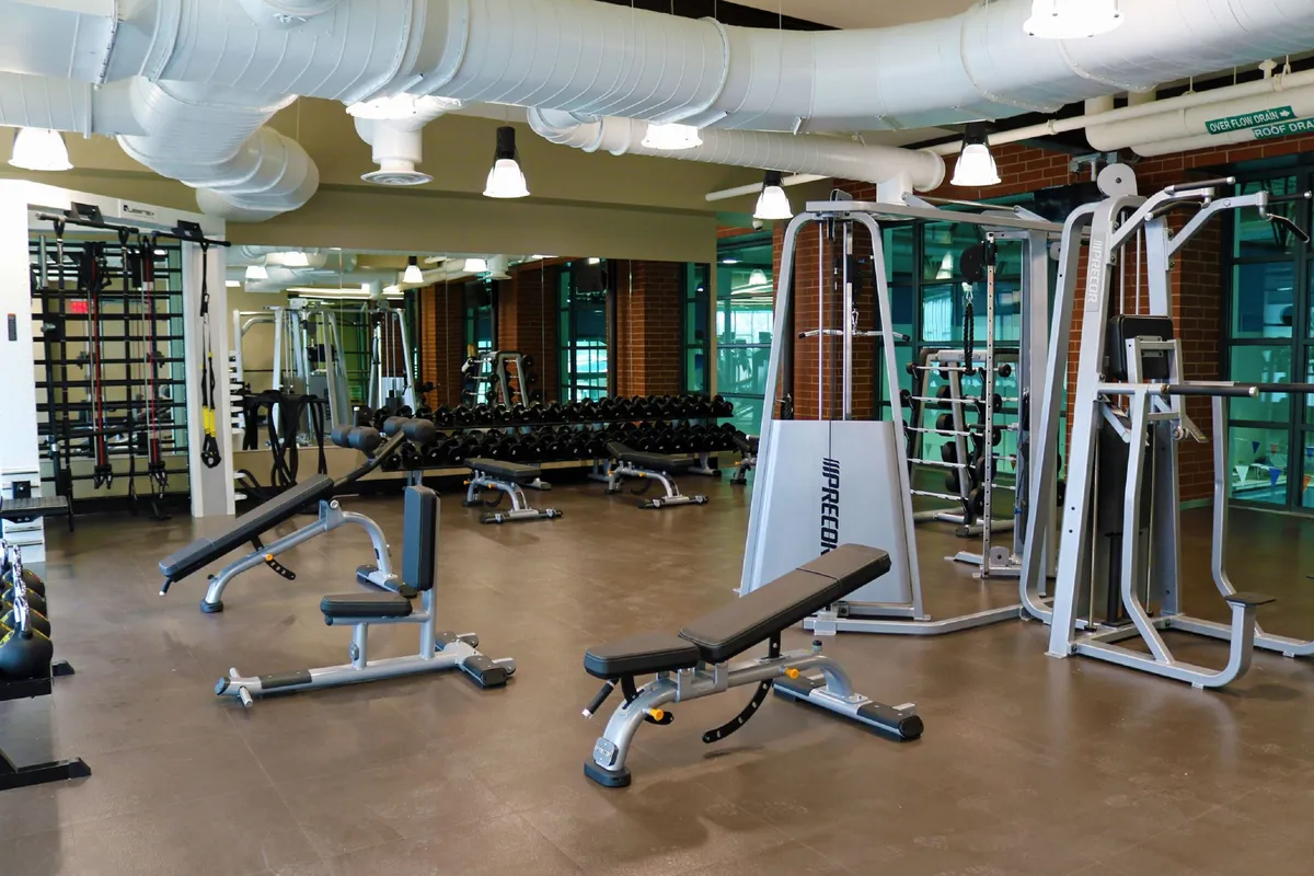 A well-equipped fitness center with a variety of exercise machines, benches, and a dumbbell rack. The bright, motivating setting suggests opportunities for personal fitness and group workouts.