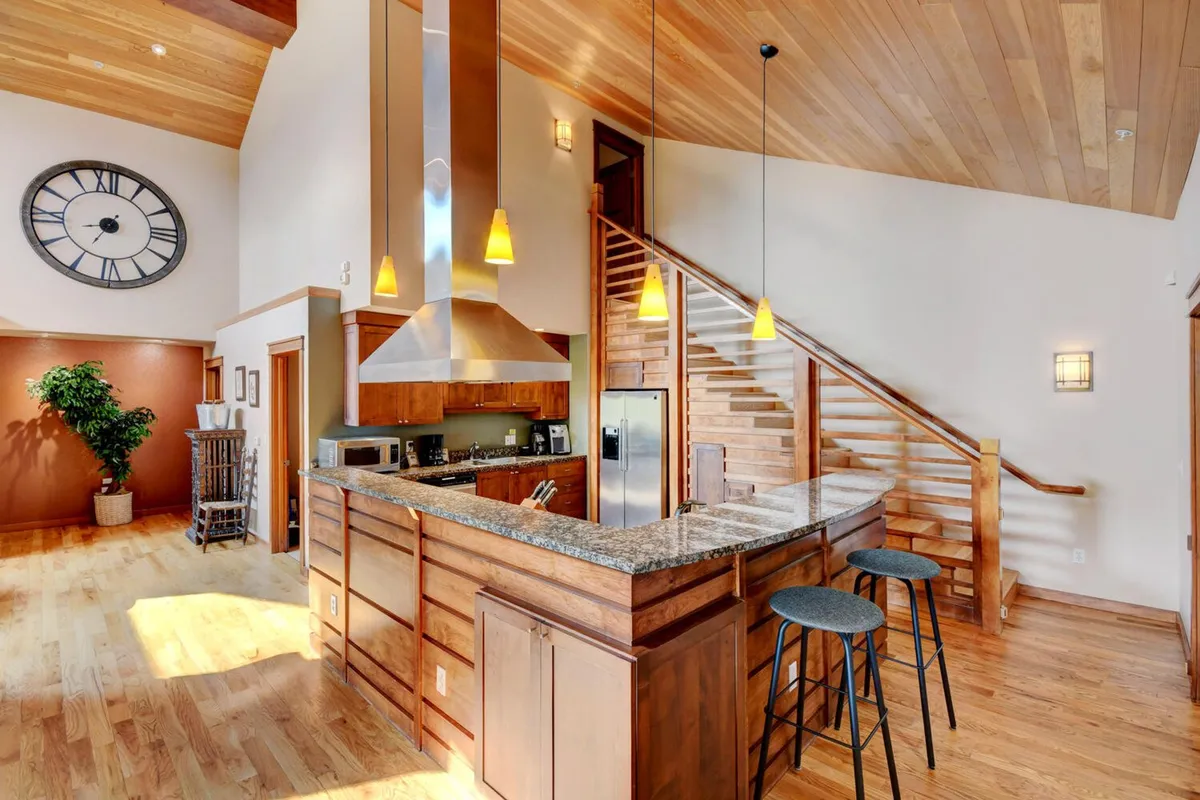 The image showcases a spacious, modern kitchen with a large island perfect for gathering, gleaming stainless steel appliances, a stylish wall clock, and an inviting staircase leading to the home's upper level. The scene exudes a warm, welcoming atmosphere ideal for entertaining, cooking, and sharing memorable moments with family or friends.
