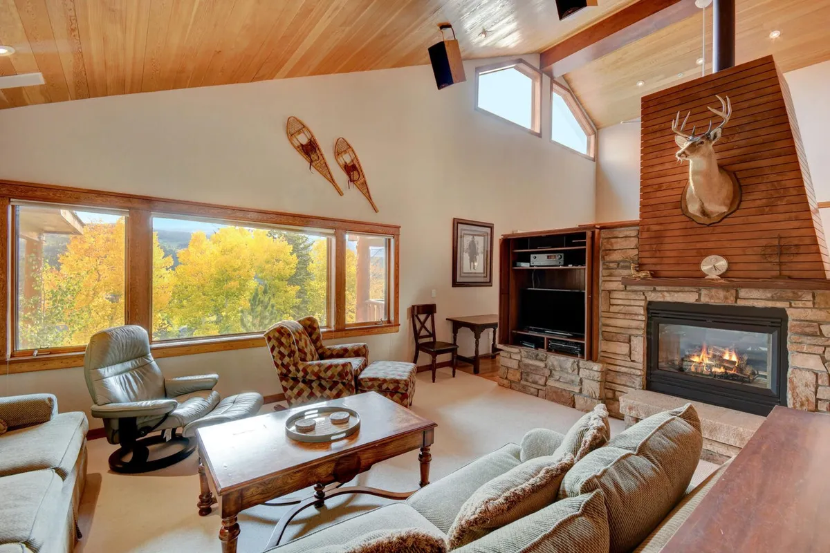 A spacious living room with a cozy fireplace, large windows showcasing an autumn mountain view, exposed wooden beams, and comfortable seating arranged for conversation or relaxation. The decor features a deer mount, evoking a refined mountain lodge atmosphere.