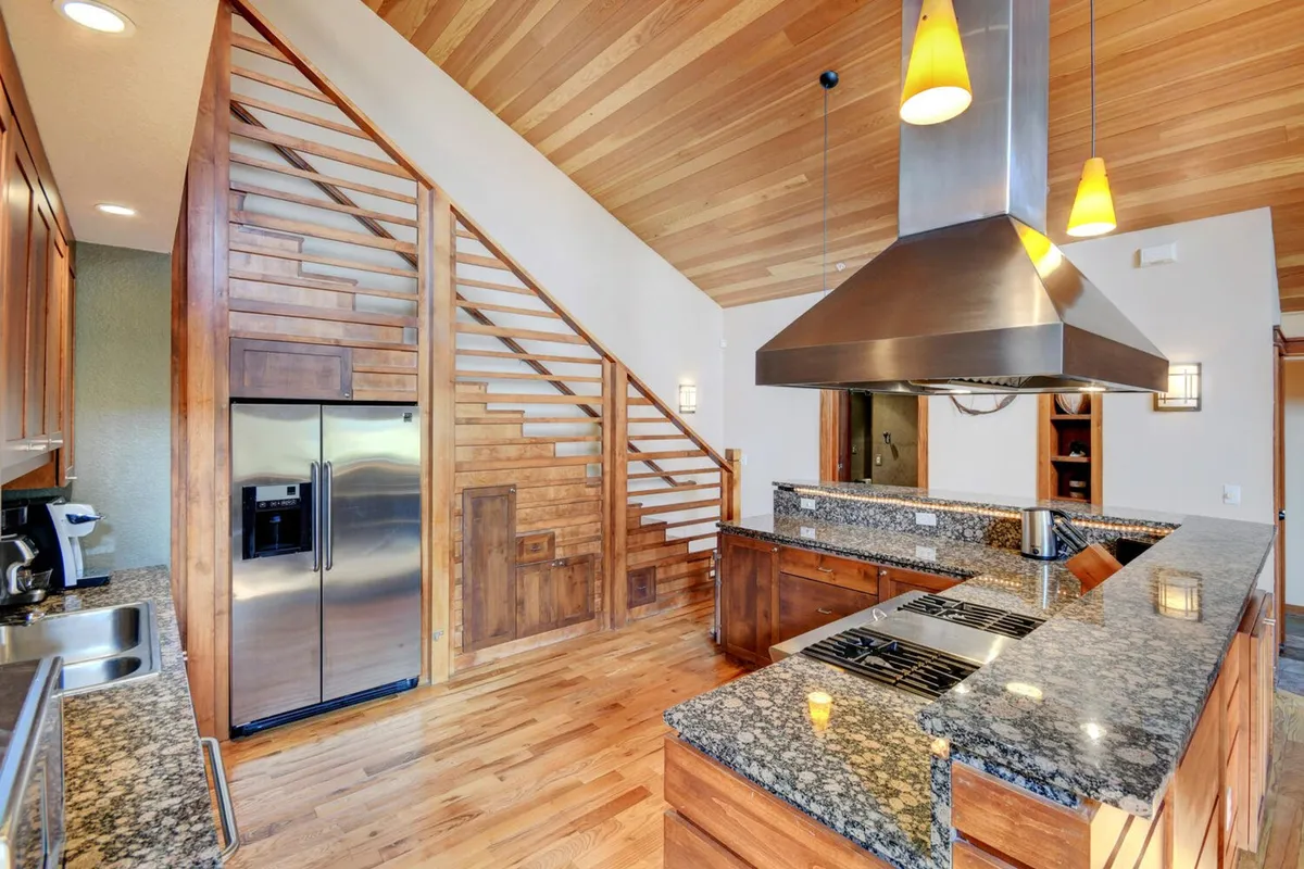 A chef’s kitchen with striking granite countertops, modern stainless appliances, rich wooden cabinetry, and a wooden staircase nearby. The space is illuminated and designed for cooking, entertaining, and gathering, with ample room for gourmet meal preparation and socializing.