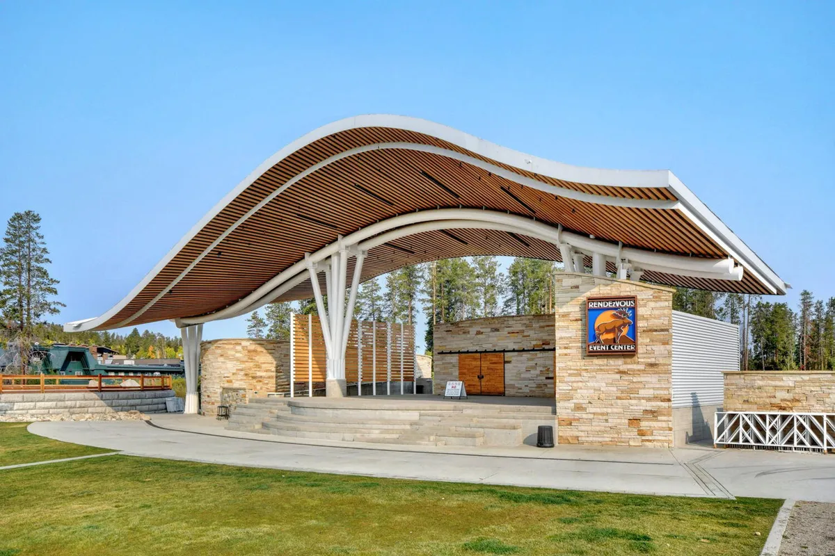 The exterior of an event center with a distinctive curved wooden roof and stone facade, showcasing signage for community events. The space is designed for open-air performances and gatherings, evoking a celebratory and communal atmosphere.